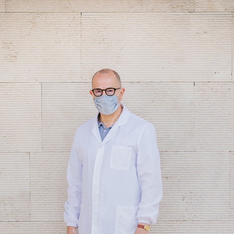A man in a white coat wearing the light blue AV Mask surgical mask against a pale stone wall.